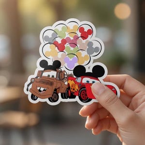 May include: A sticker featuring two cartoon cars, one brown and one red, with Mickey Mouse ears. Above the cars are colourful Mickey Mouse head balloons. The sticker is held by a hand.
