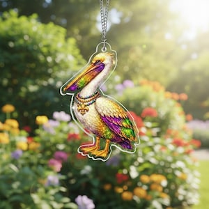 Op de afbeelding: Een heldere acryl hanger met een kleurrijk pelikaan ontwerp. De vogel heeft een gele kop, een wit lichaam en iriserende vleugels met paarse, groene en gouden accenten. Het draagt een kralenketting met een fleur-de-lis hanger en hangt aan een zilveren ketting.