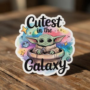 Op de afbeelding: Een gestanste sticker met een groen, buitenaards personage in een ronde mand, met de tekst "Cutest in the Galaxy". De achtergrond is een aquarel wassing van blauw, roze en paars met sterren en een klein wezen.