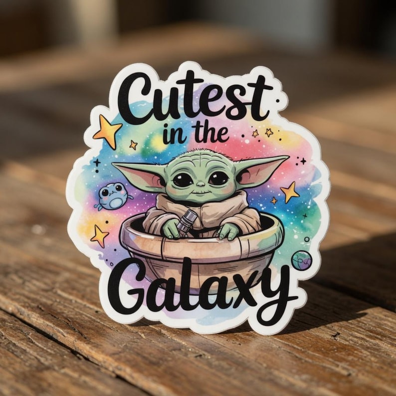 Op de afbeelding: Een gestanste sticker met een groen, buitenaards personage in een ronde mand, met de tekst "Cutest in the Galaxy". De achtergrond is een aquarel wassing van blauw, roze en paars met sterren en een klein wezen.