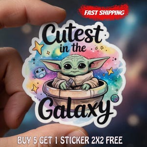 Op de afbeelding: Een kleurrijke sticker met een stripfiguur in een zwevende capsule. De sticker heeft de tekst "Cutest in the Galaxy" met sterren en een klein wezen. De woorden "Fast Shipping" zijn ook zichtbaar.