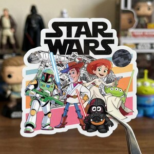Puede incluir: Una colorida pegatina con personajes de Star Wars y Toy Story. El logotipo de Star Wars est&aacute; en la parte superior. Los personajes incluyen a Boba Fett, Woody, Jessie, Darth Vader y otros, con un fondo de nave espacial y planeta.