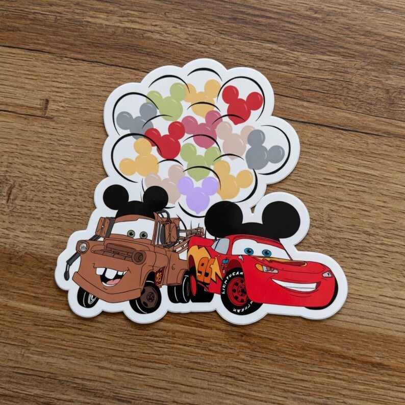 May include: A die-cut sticker featuring two cartoon cars, one brown and one red, with Mickey Mouse ears. Above them are colorful Mickey Mouse head shapes. The sticker is on a wooden surface.