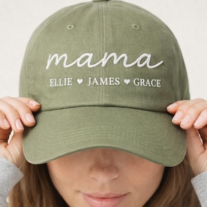 May include: Olive green baseball cap with the word "mama" embroidered in white script. Below the word "mama" are the names "Ellie", "James", and "Grace", each separated by a heart symbol. The cap is being held by a person.