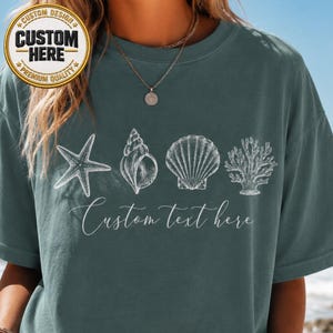 May include: A dark teal crewneck t-shirt featuring a white graphic print of a starfish, a spiral seashell, a fan-shaped scallop shell, and a piece of coral. Below the marine life illustrations, the text Custom text here is printed in white.