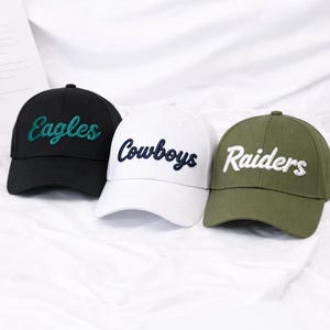 May include: Three baseball caps in black, white, and olive green. Each cap features embroidered text: "Eagles" in teal, "Cowboys" in navy, and "Raiders" in white. The caps have a curved brim and a classic, casual style.