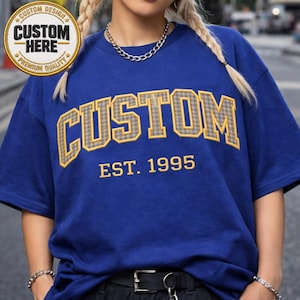 May include: A royal blue t-shirt with the word "CUSTOM" in a yellow and gray plaid design, with "EST. 1995" below. A circular emblem in the upper left corner reads "CUSTOM HERE" and "PREMIUM QUALITY".