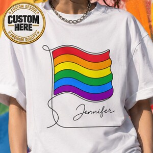May include: White t-shirt featuring a rainbow flag design with the name "Jennifer" in cursive. The flag has red, orange, yellow, green, blue, and purple stripes. The shirt has a round neck and short sleeves.