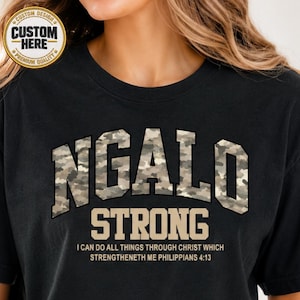 May include: A black t-shirt featuring the word NGALO in a camouflage pattern, with the word STRONG and the text I CAN DO ALL THINGS THROUGH CHRIST WHICH STRENGTHENETH ME PHILIPPIANS 4:13 printed in a tan color below the main graphic.