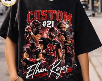 May include: A black t-shirt featuring a graphic print of a football player wearing a black and red uniform with the number 21. The design includes the text CUSTOM #21 and the name Ethan Reyes in white script, surrounded by red lightning effects.