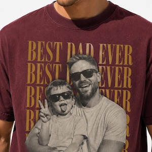 May include: Burgundy t-shirt with the words "BEST DAD EVER" in gold lettering. The shirt features a black and white photo of a man and child wearing sunglasses. The child is sticking out their tongue.