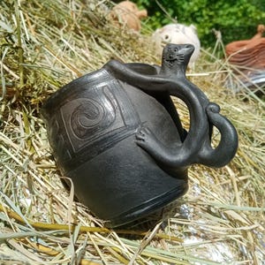 May include: A handmade black ceramic mug with a unique handle shaped like a creature. The mug features carved spiral designs and is set against a backdrop of hay and greenery. The handle has a detailed face and body.