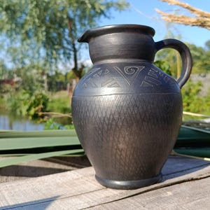 Könnte beinhalten: Ein schwarzer Keramikkrug mit Henkel und Ausguss, verziert mit geätzten geometrischen Mustern. Der Krug steht auf einer Holzoberfläche mit einem natürlichen Hintergrund aus Grün und blauem Himmel.