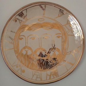 May include: A cream-coloured ceramic plate with a gold rim and a central design of three faces with halos and beards. The faces are rendered in gold, with additional gold detailing around the edges of the plate.