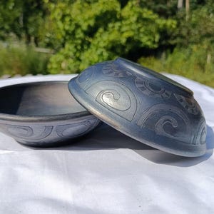 May include: Two dark grey ceramic bowls with etched patterns. One bowl is upright, the other is tilted. The bowls are on a white surface, with a blurred green background of trees and foliage.