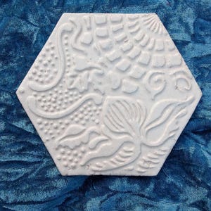 Puede incluir: Una baldosa hexagonal blanca con un diseño floral y abstracto en relieve. La baldosa presenta un patrón detallado de hojas, espirales y líneas punteadas. La baldosa está colocada sobre un fondo azul oscuro texturizado.