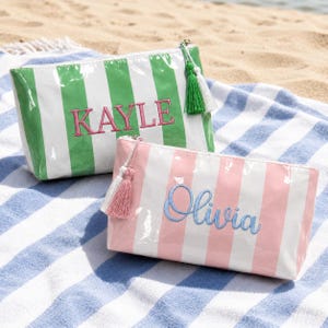 Puede incluir: Dos estuches rectangulares con cremallera y acabado brillante. Uno presenta rayas verticales verdes y blancas con el nombre KAYLE bordado en rosa. El otro tiene rayas verticales rosas y blancas con el nombre Olivia bordado en azul.