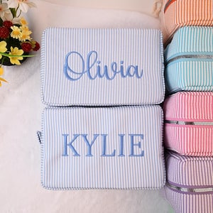 Può includere: Due trousse rettangolari a righe, una con "Olivia" ricamato in blu, l'altra con "Kylie". Ulteriori borse in arancione, verde acqua, rosa e viola sono impilate a destra. Le borse hanno una chiusura con cerniera.