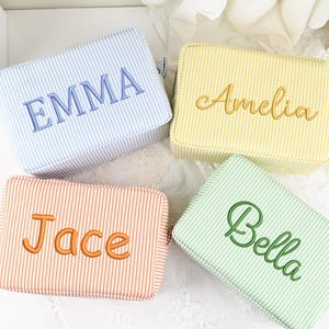 Puede incluir: Cuatro neceseres rectangulares a rayas en colores pastel: azul con "EMMA", amarillo con "Amelia", melocotón con "Jace" y verde con "Bella". Cada neceser tiene un nombre bordado.