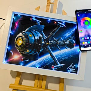 Könnte beinhalten: Ein gerahmtes, beleuchtetes Kunstwerk mit einem detaillierten Raumschiffdesign vor dem Hintergrund von Weltraum und Planeten. Das Kunstwerk befindet sich in einem weißen Rahmen mit LED-Leuchten. Ein Smartphone zeigt eine Farbregelungsoberfläche. Das Kunstwerk steht auf einer Holzstaffelei.