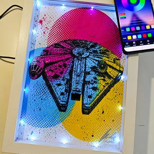 Könnte beinhalten: Ein gerahmtes Kunstwerk mit einem farbenfrohen Millennium Falcon-Design. Das Kunstwerk wird von LED-Leuchten beleuchtet und vor einem Hintergrund aus rosa, blauen und gelben Kreisen platziert. Ein Smartphone zeigt eine Farbsteuerungs-App.
