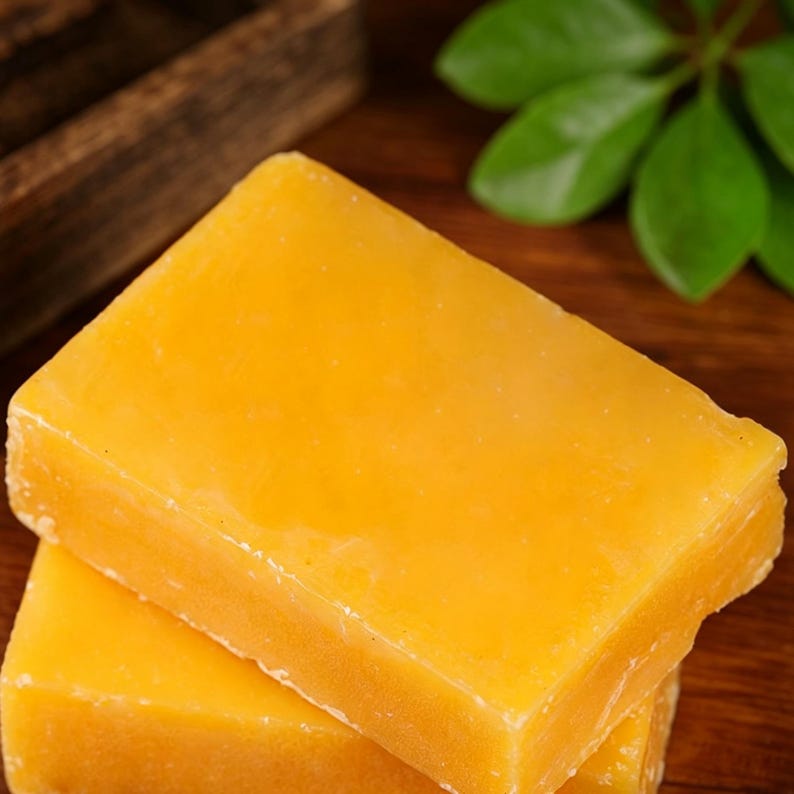 May include: Two rectangular blocks of yellow cheese are stacked on top of each other. The cheese has a smooth, slightly glossy surface. The background includes a wooden box and green leaves.