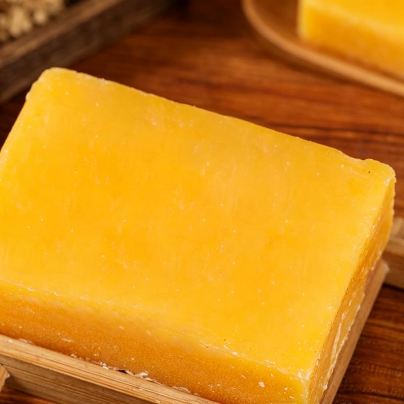 May include: A close-up shot of a block of cheddar cheese. The cheese is a vibrant yellow-orange color and has a slightly rough texture. It sits on a wooden surface, suggesting a rustic or artisanal presentation.