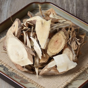 May include: A close-up shot of a square ceramic dish filled with dried, light brown herbal slices and pieces. The pieces vary in size and shape, with some showing the cross-section of a stem. The dish sits on a burlap mat.