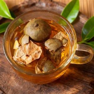 May include: A clear glass teacup filled with amber-colored tea and several dried, brown, spherical and petal-shaped tea leaves. The cup sits on a wooden surface, with green leaves in the background.