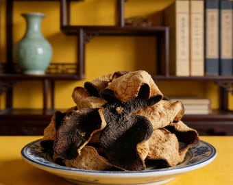May include: A pile of dried citrus peels, some dark brown and others tan, are arranged on a white and blue patterned plate. The background features a yellow wall, a green vase, and a dark wooden shelf with books.