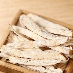 Może przedstawiać: Zbliżenie suszonych, jasno zabarwionych, pokrojonych korzeni ułożonych w drewnianej tacy. Korzenie mają lekko szorstką teksturę i blado-beżowy odcień, z kilkoma ciemniejszymi plamami. Drewniana taca ma naturalny, jasnobrązowy kolor.