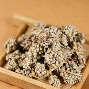 Può includere: Primo piano di boccioli di erbe secche color beige in un piccolo vassoio di legno marrone chiaro. I boccioli hanno un aspetto strutturato e raggruppato. Lo sfondo è un marrone caldo sfocato.