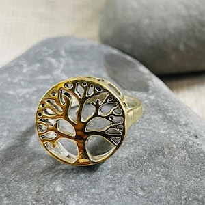 Puede incluir: Un anillo dorado con un diseño de árbol de la vida. El anillo presenta un marco circular con una silueta detallada de un árbol. El anillo está colocado sobre una piedra gris, con una segunda piedra en el fondo.