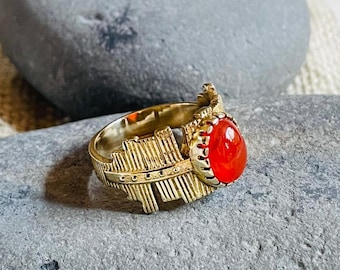 May include: A gold-colored ring with an oval, bright red gemstone. The ring has a detailed design with a feather-like pattern on the band. The ring is displayed on a gray stone, with a neutral background.