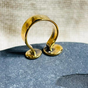 May include: A gold-colored ring with a unique design. The ring features a curved band that rises from two small, circular bases. The ring is displayed on a gray stone, showcasing its shape and metallic sheen.