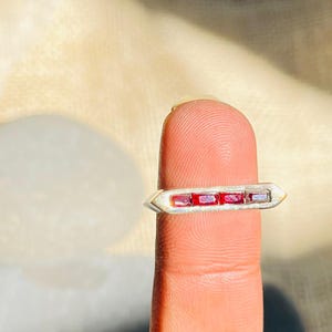 Peut inclure: Une bague en argent ornée de trois pierres rouges rectangulaires disposées en ligne. La bague présente un design simple et moderne, avec des extrémités légèrement pointues. La bague est présentée sur un doigt.