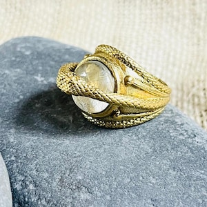 以下が含まれることがあります： 金色のリングで、透明で丸い宝石が特徴です。リングのバンドは、石の周りに巻き付く蛇のモチーフで複雑にデザインされています。リングは灰色の石の上に展示されており、ニュートラルな生地の背景があります。