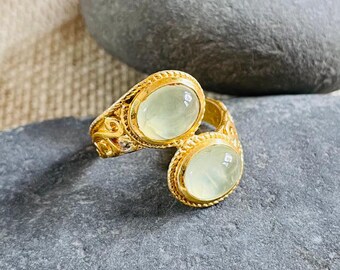 Può includere: Un anello color oro con due pietre preziose ovali verde pallido. L'anello ha un design ornato e strutturato attorno alle pietre preziose e sulla fascia. L'anello è esposto su una superficie di pietra grigia.