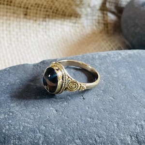 May include: A gold-colored ring with an oval-shaped, dark-colored stone. The ring has intricate detailing around the stone and on the band. The ring is displayed on a gray stone, showcasing its design and craftsmanship.