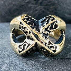 Puede incluir: Un anillo dorado con un diseño de símbolo de infinito. El anillo tiene un patrón intrincado y texturizado y un acabado pulido. El anillo está sobre un fondo de piedra gris.