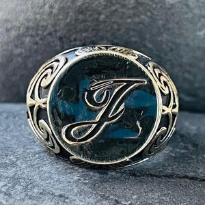 May include: Silver-toned ring with a black background and the letter "J" in a cursive font. The ring has an ornate, swirling design around the letter and a dark blue center. The ring is set on a gray stone surface.