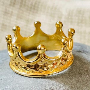 Può includere: Un anello a corona dorata con superficie strutturata. L'anello presenta un design a corona classico con più punte e dettagli arrotondati. L'anello è appoggiato su una pietra grigia, con uno sfondo neutro.