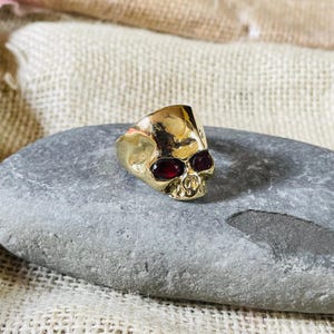 May include: A gold-colored skull ring with red gemstone eyes. The ring is set on a gray stone surface. The skull design features detailed facial features, including eye sockets, a nose, and teeth. The ring is a unique piece of jewelry.