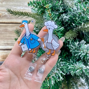 Può includere: Due porta badge in acrilico con oche dei cartoni animati. Uno è un medico con un camice blu, l'altro è uno chef con un cappello e un grembiule bianchi. Entrambi hanno becchi e zampe arancioni. I porta badge hanno clip in plastica trasparente.