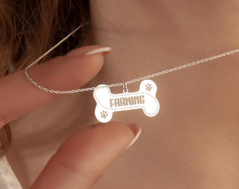 May include: A silver-colored necklace with a bone-shaped pendant. The pendant has the word "FARMING" engraved in the center, with paw prints on either side. The necklace is being held up by a person.