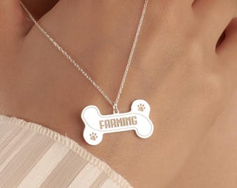 May include: A silver-colored dog bone pendant necklace. The bone-shaped pendant has the word "FARMING" engraved in the center, with paw prints on each end. The necklace is on a delicate chain.