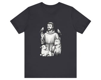 May include: Dark gray t-shirt featuring a black and white illustration of a saint with birds, a lamb, and a wolf. The saint is depicted with a halo, wearing a robe, and has a beard. The design is centered on the shirt.