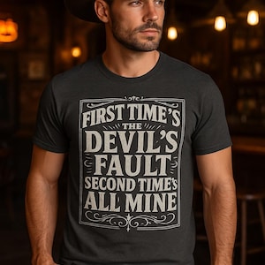 Puede incluir: Camiseta gris oscuro con el texto blanco "FIRST TIME'S THE DEVIL'S FAULT SECOND TIME'S ALL MINE" en un diseño vintage. La persona de la imagen lleva un sombrero de vaquero marrón y vaqueros azules.