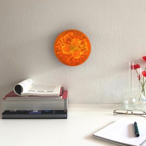 Puede incluir: Una obra de arte mural circular de color naranja y amarillo con un diseño texturizado, montada en una pared blanca. La imagen también muestra una pila de libros, un cuaderno y un jarrón con flores rojas. La obra de arte se asemeja a un sol.