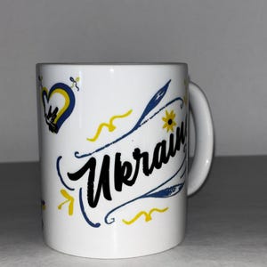 May include: White ceramic mug with the word "Ukraine" in black script, accented with blue and yellow floral and heart designs. The mug has a white handle and is likely used for hot beverages.